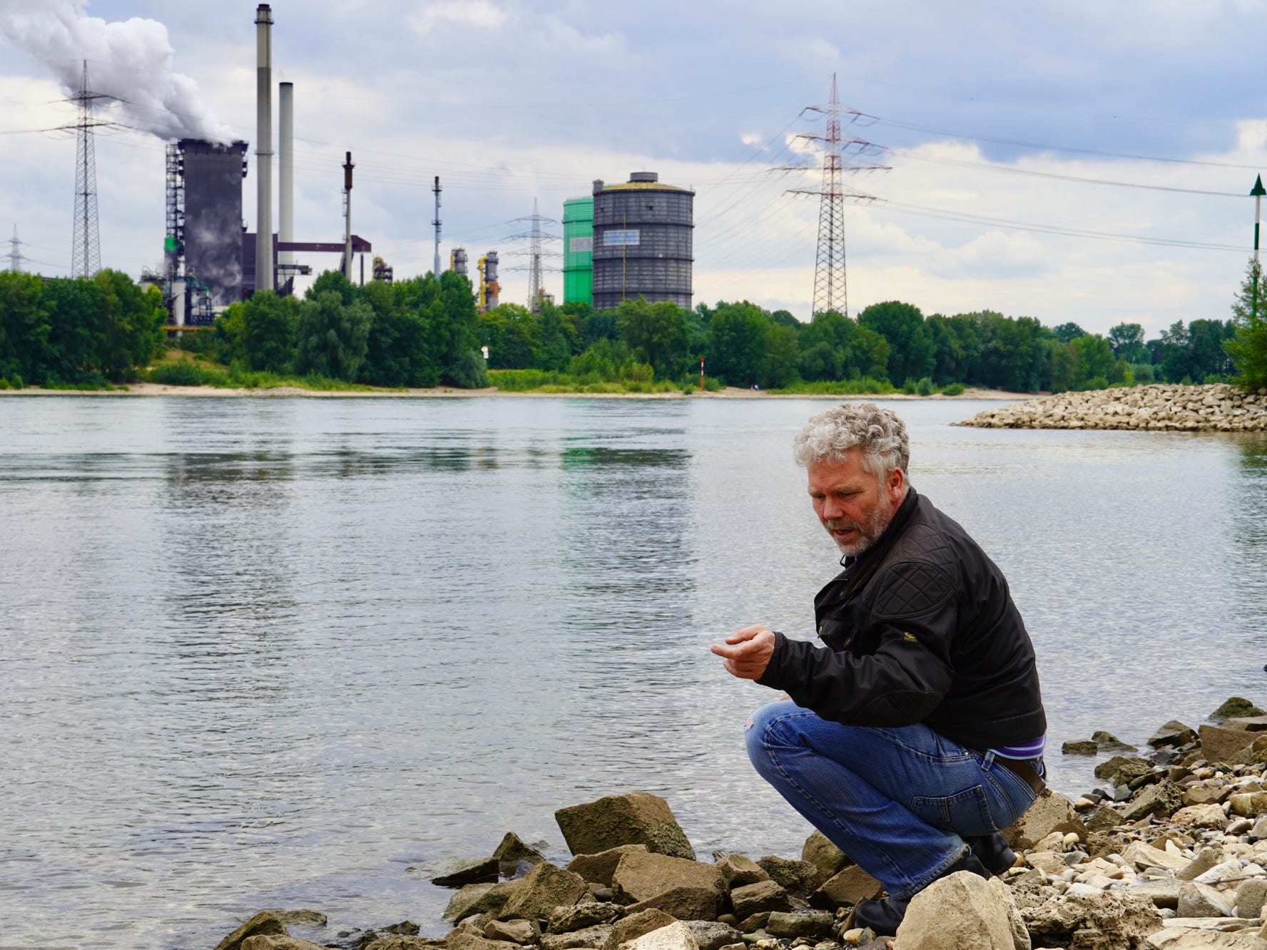 Stephan Schütze hockt direkt am Rhein bei Duisburg und betrachtet den Zustand des Ufers.